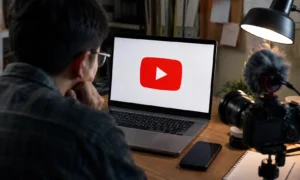 A content creator sits at a desk looking at a laptop displaying the YouTube logo, with a camera, microphone, smartphone and notebook nearby in a home studio setup.